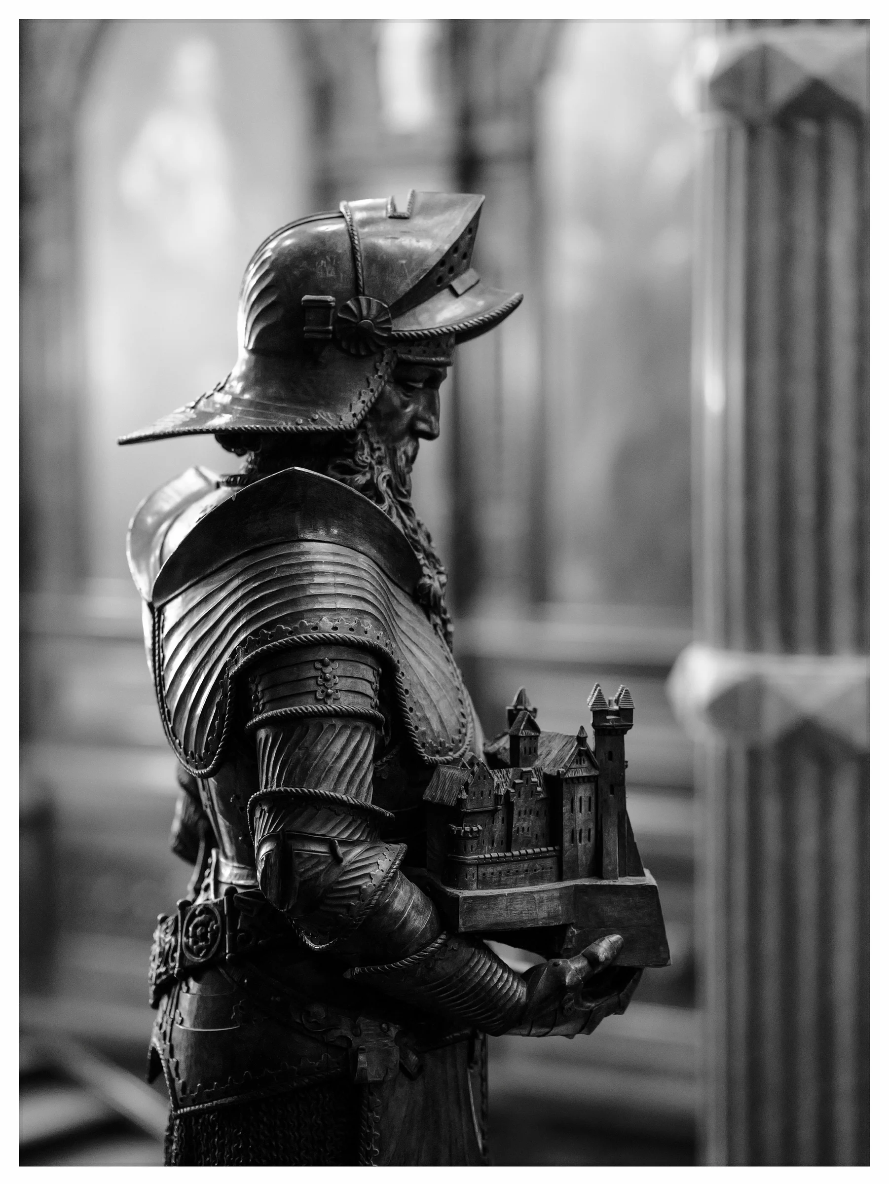 Black and white image of an armour-clad statue holding a miniature model of Peleș Castle