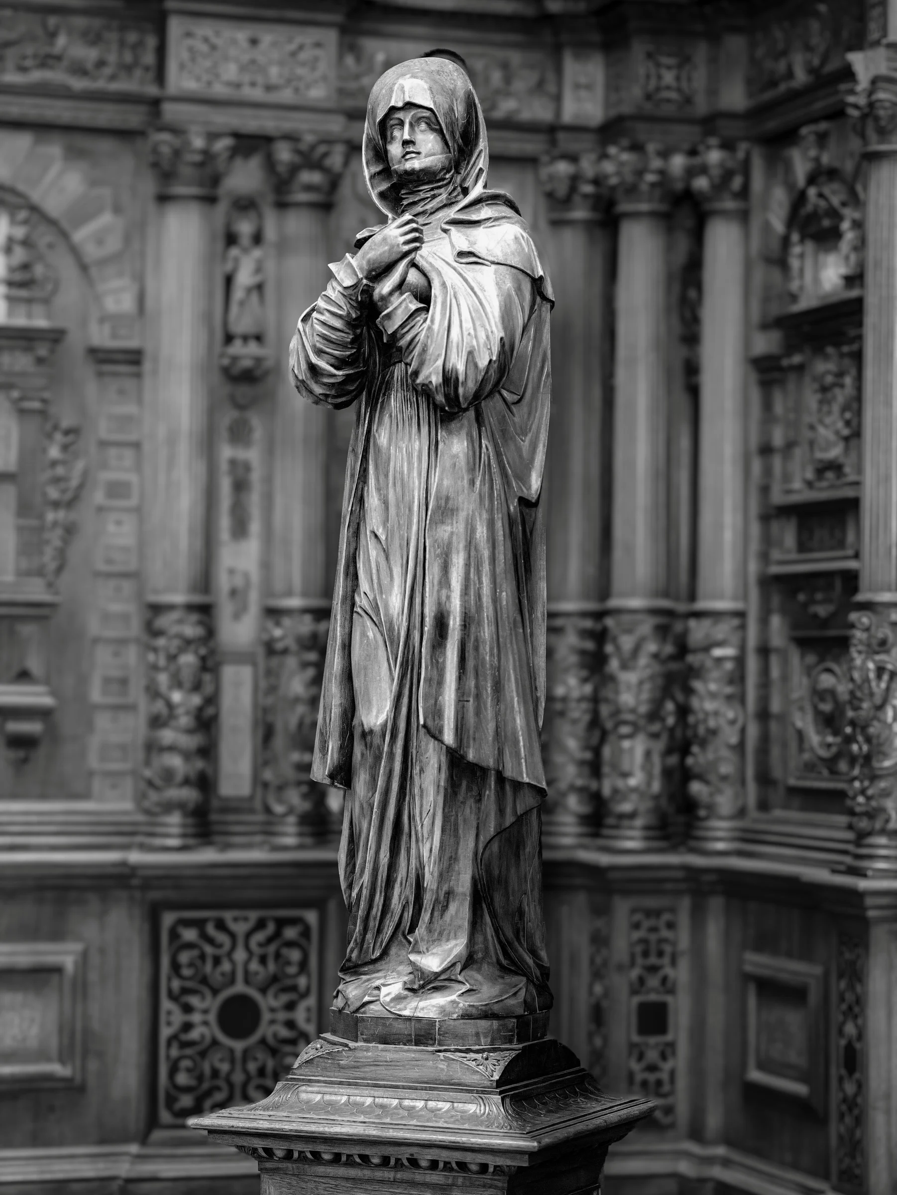Black and white photo of a solemn statue draped in a cloak standing on a detailed pedestal inside Peleș Castle, Romania, with intricate carved stone walls in the background.