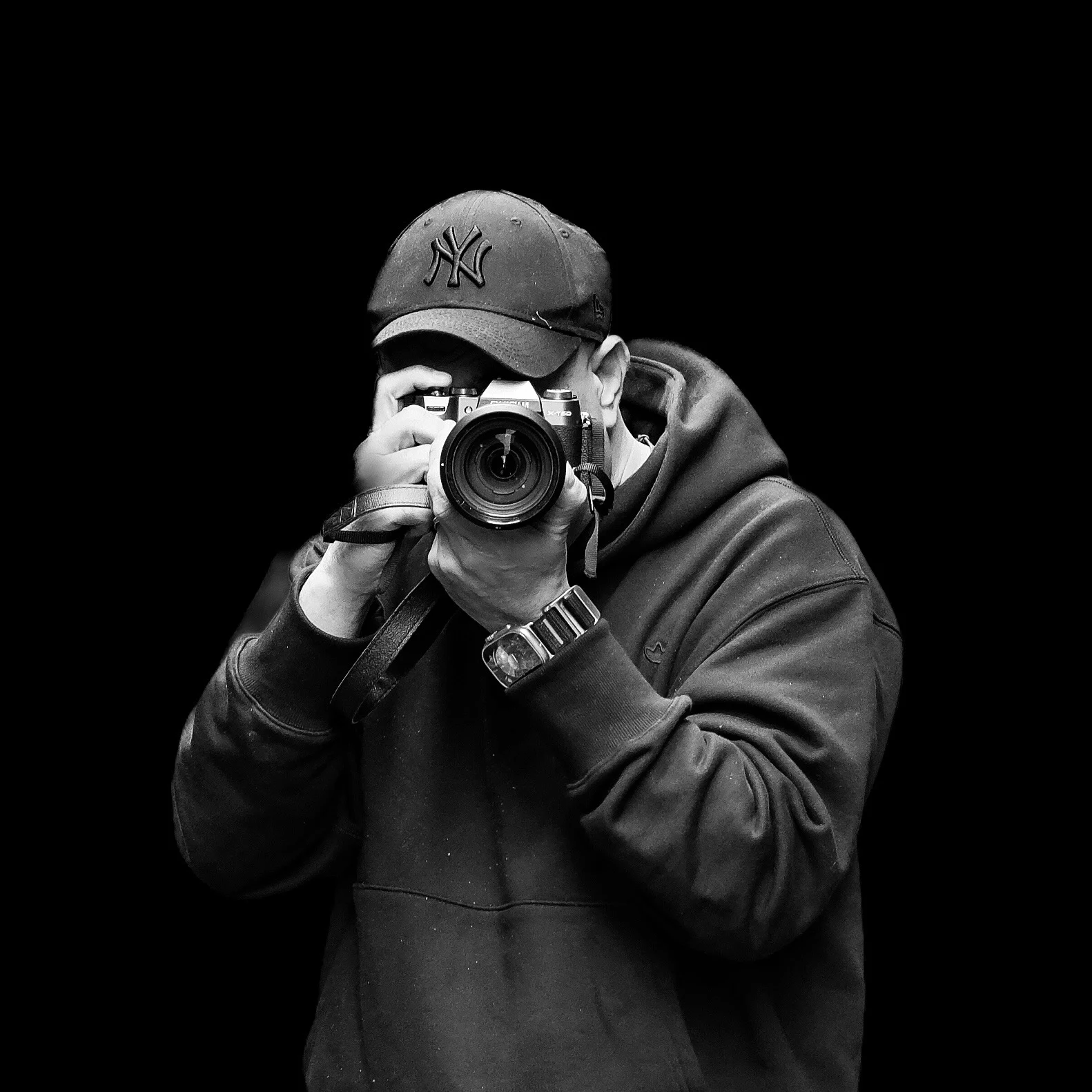 Black and white photo of a person holding a camera up to their face, wearing a hooded sweatshirt and a cap