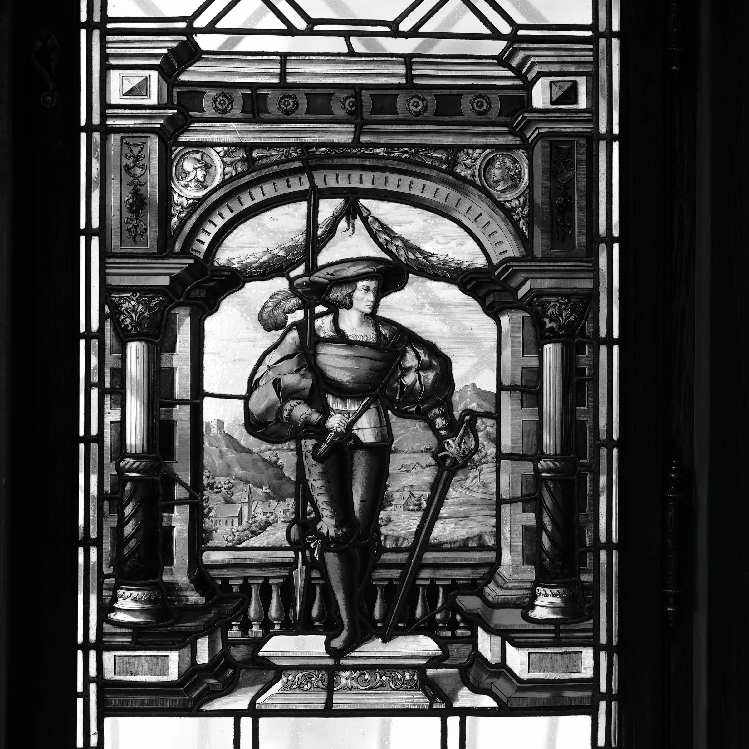 Black and white stained glass window depicting a figure in armor holding a sword with a mountainous landscape background