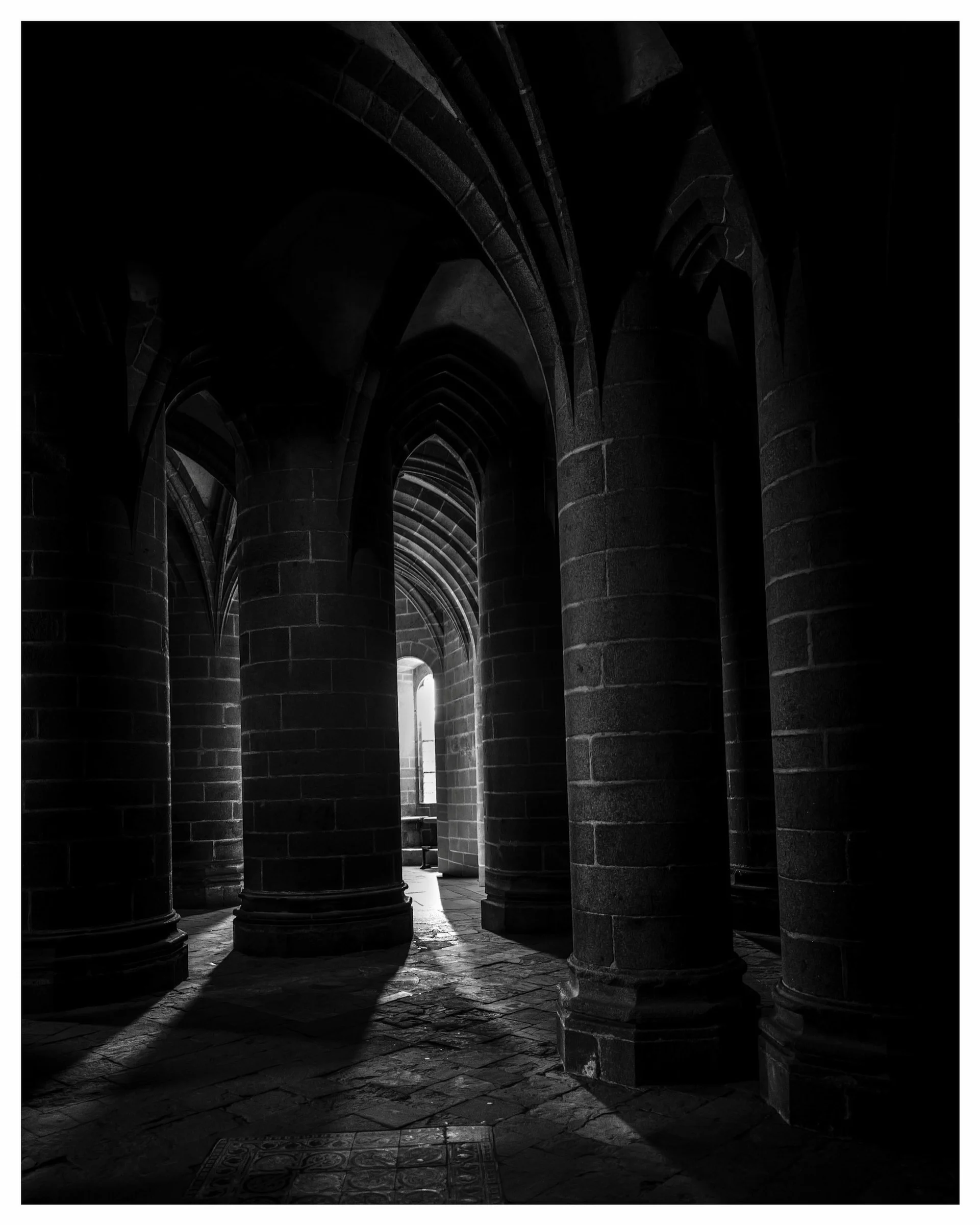 Black and white photo of stone pillars and arches inside Mont Saint Michel abbey with light streaming through a narrow opening