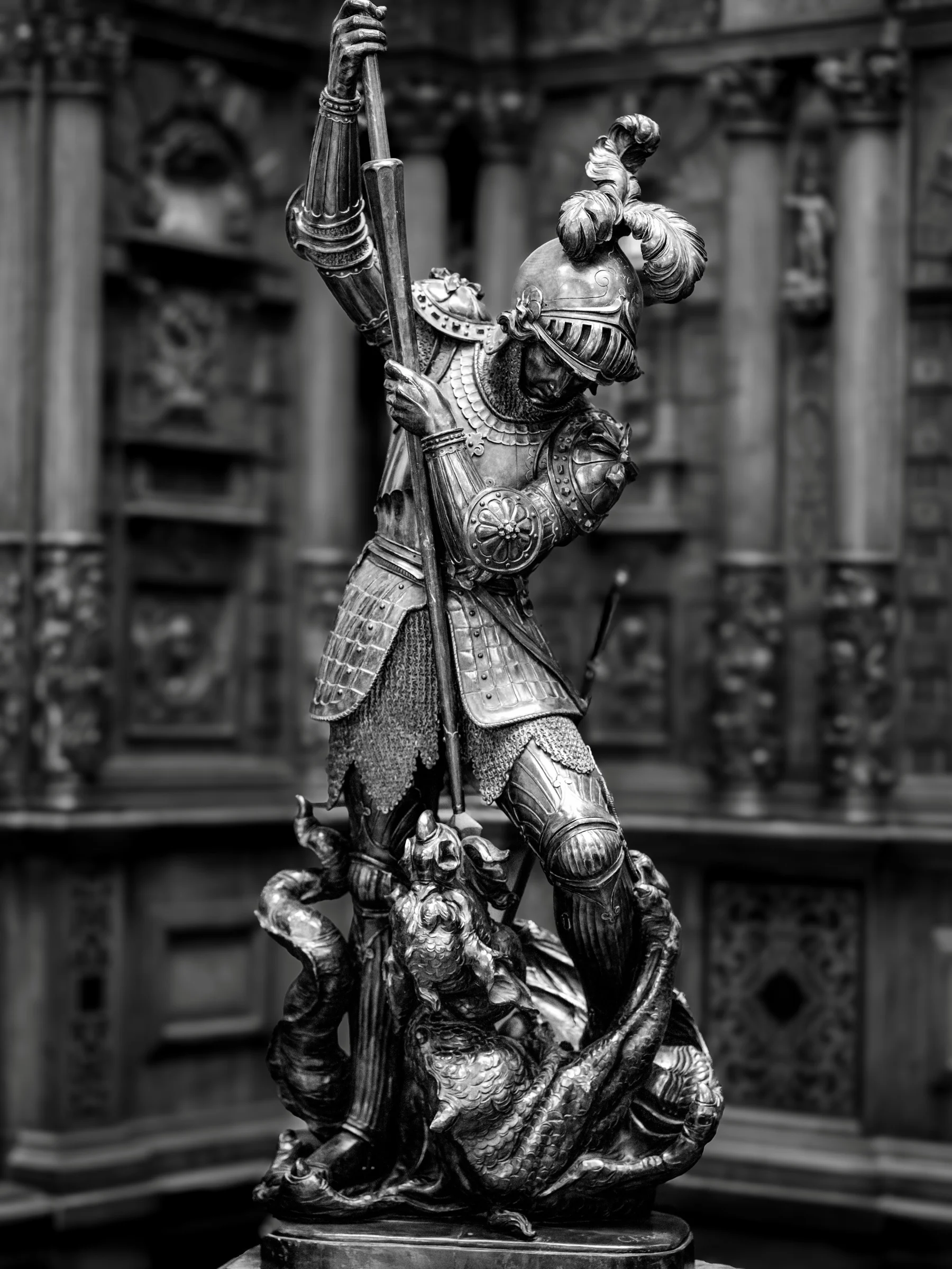 Black and white photograph of a detailed wooden sculpture of Saint George in armor, raising a spear above a dragon, inside Peleș Castle, Romania.