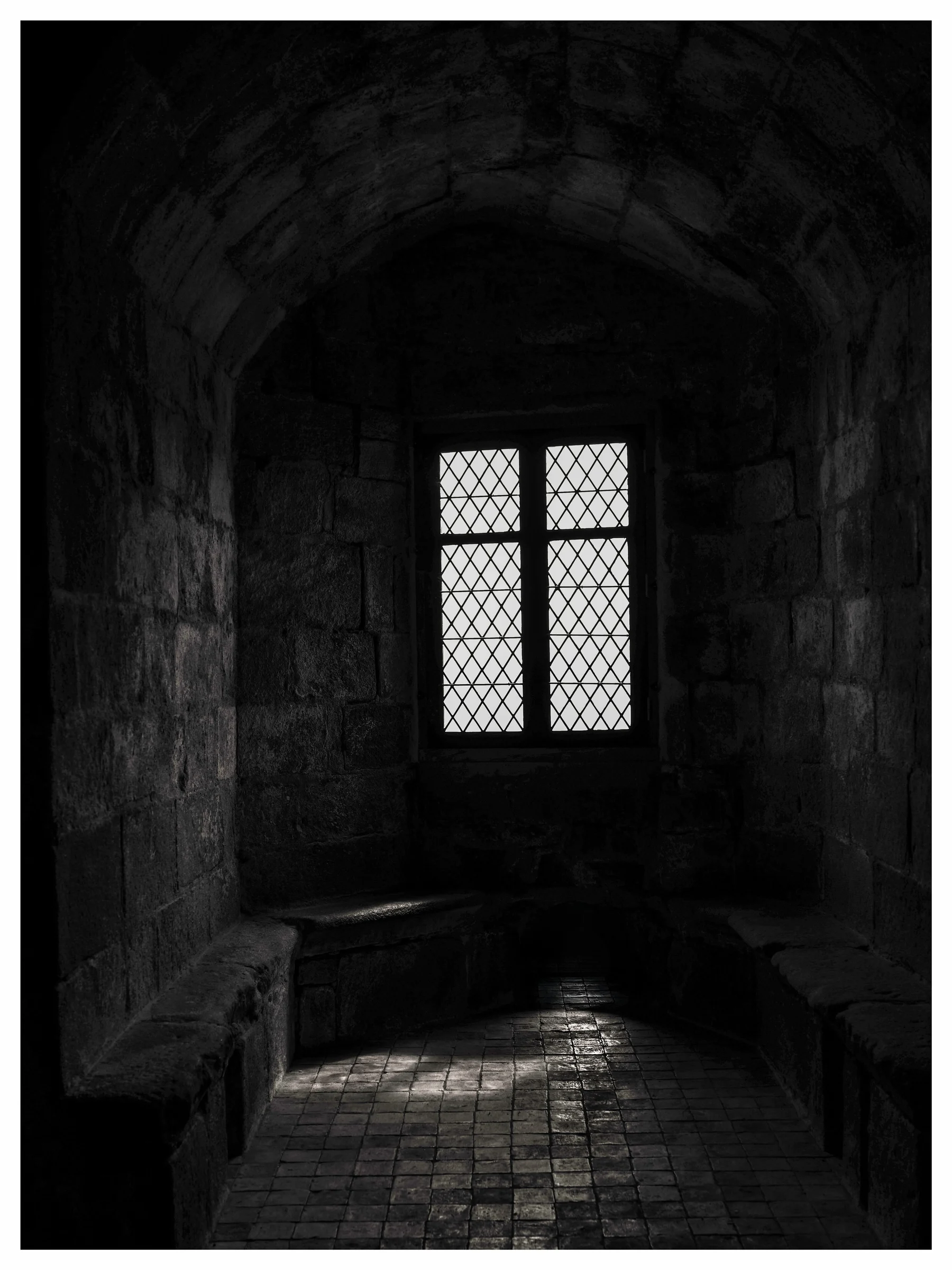 Black and white photo of a small window in a dark stone room with light on the tiled floor