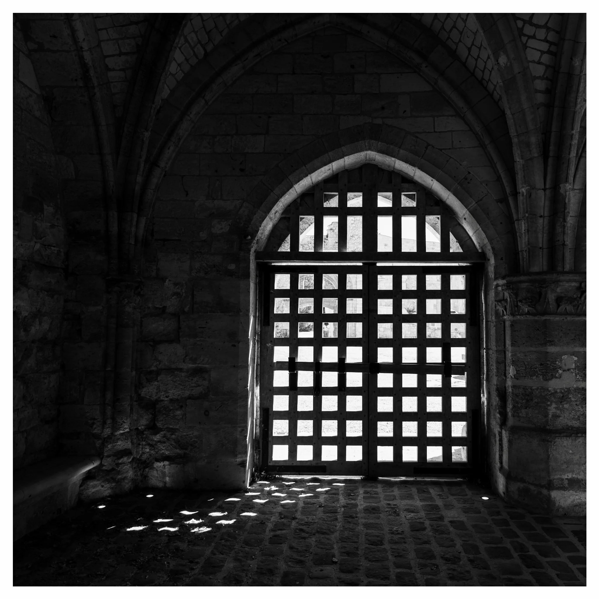 Interior view of a stone arched doorway with a heavy grid-patterned gate casting light shadows on a cobblestone floor in a historic stone building.